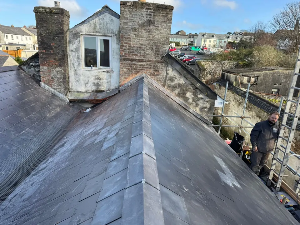 Finished slate roof with a new hip beside a dormer and two brick chimneys, roofer on the scaffold